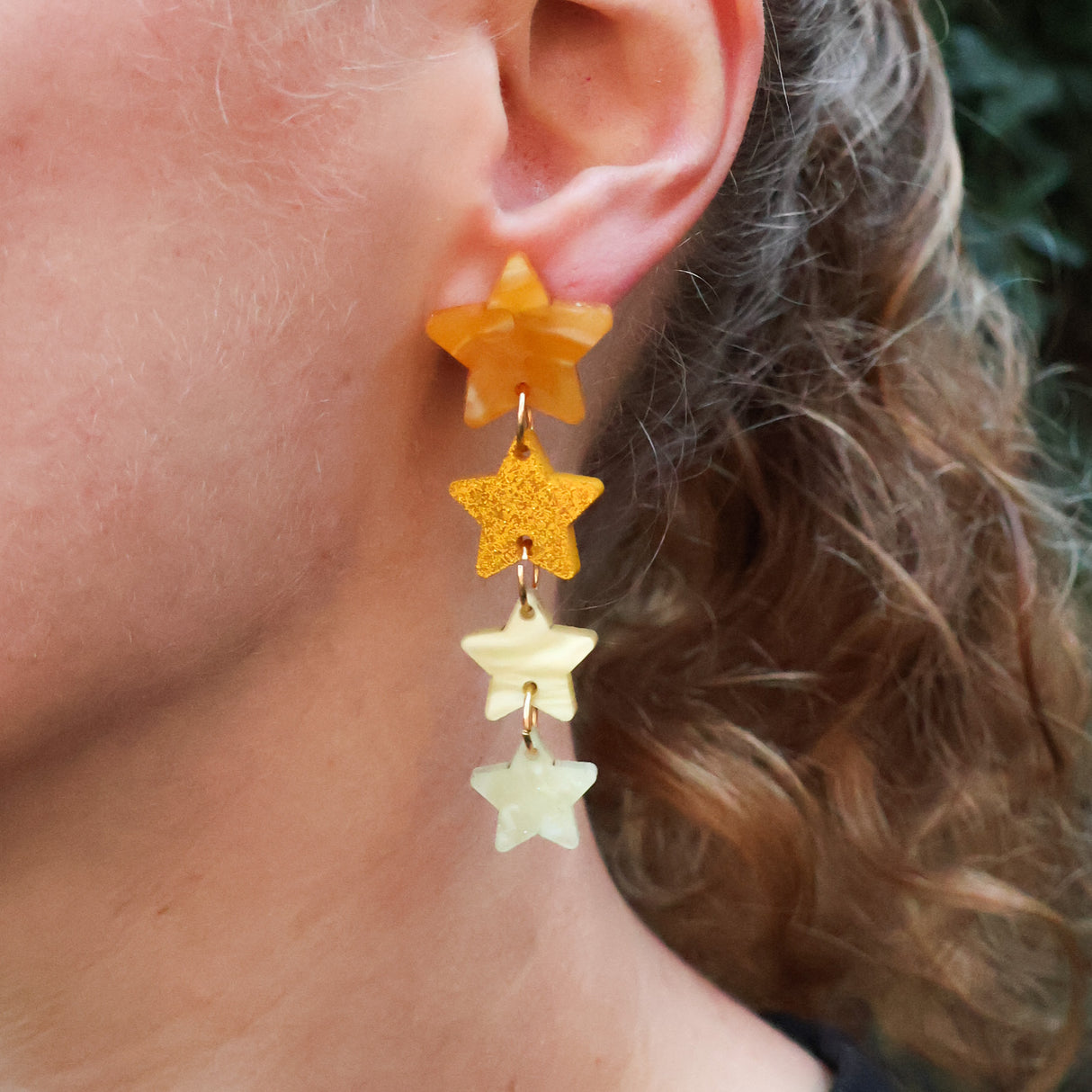 Closeup of a person wearing acrylic star earrings. 4 stars in 4 different yellow and gold toned acrylics are stacked. 