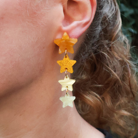 Closeup of a person wearing acrylic star earrings. 4 stars in 4 different yellow and gold toned acrylics are stacked. 
