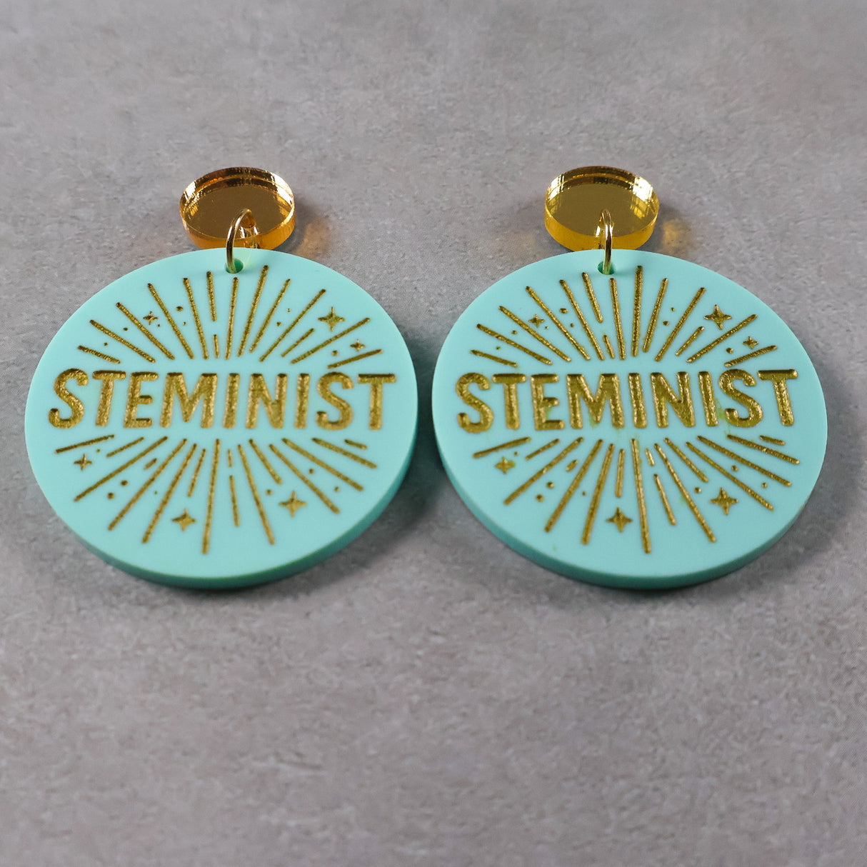 Turquoise or teal dangle earrings with 'STEMINIST' gold text and gold mirror acrylic toppers