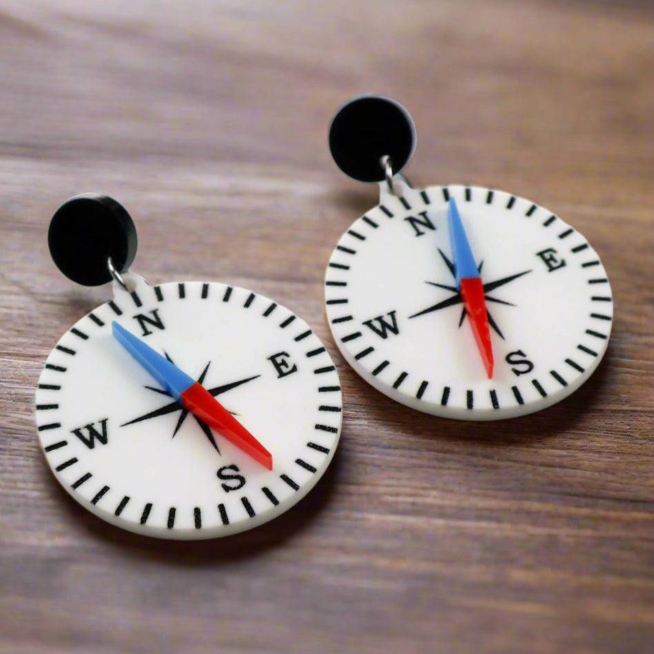 A pair of compass-themed acrylic earrings on a wooden background. The compass is white, with a red and blue needle, and round black toppers. 