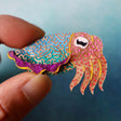 Colourful acrylic cuttlefish brooch held between fingers against a blue background