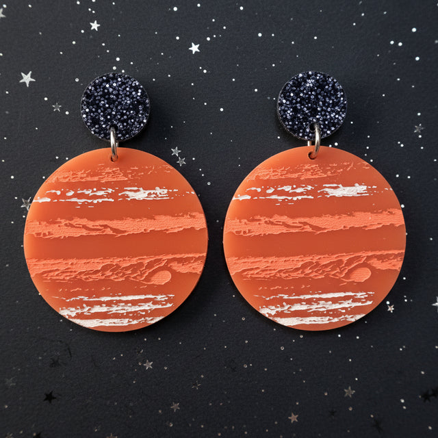 A pair of earrings with an orange jupiter design, hanging from back glitter toppers. 