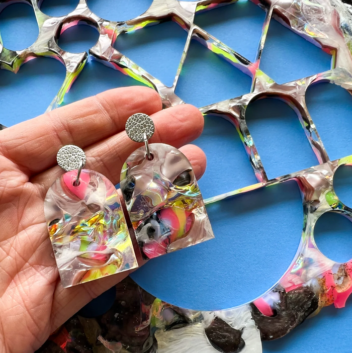 Colourful arch shaped earrings held in a hand with a blue background and the leftovers of a recycled acrylic sheet. 