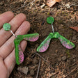 A pair of acrylic earrings shaped like maple samara helicopter seeds being held in a hand