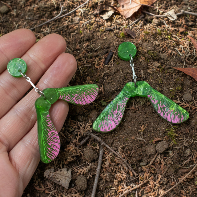 A pair of acrylic earrings shaped like maple samara helicopter seeds being held in a hand