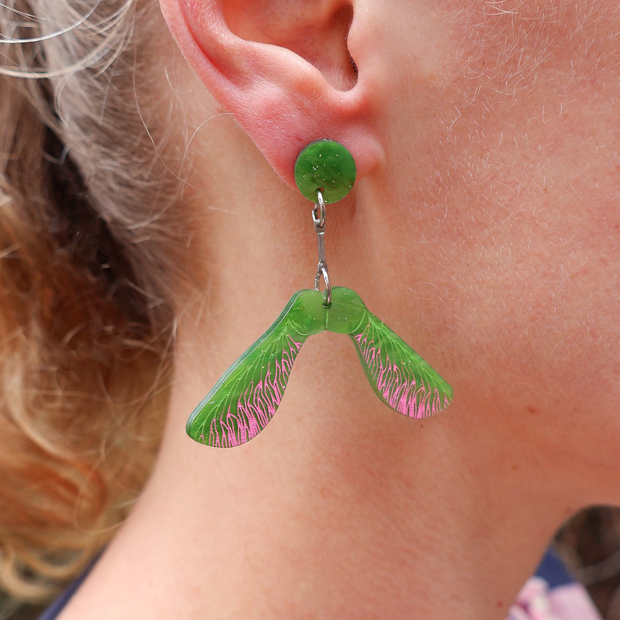 Close-up of a person wearing green acrylic earrings shaped like a maple helicopter samara seed.