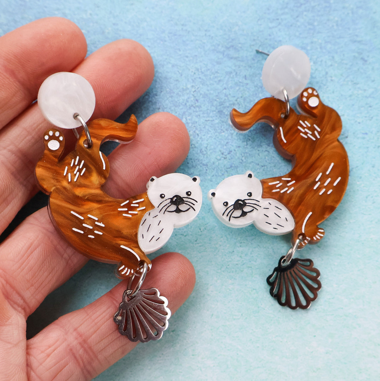 A Pair of otter-shaped earrings with a hand holding one, on a light blue background.