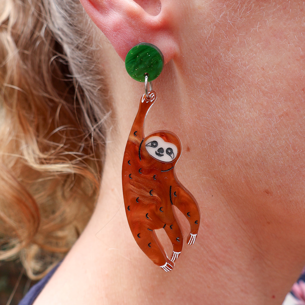 Brown Acrylic sloth earrings hanging from a green acrylic topper, being modelled. 