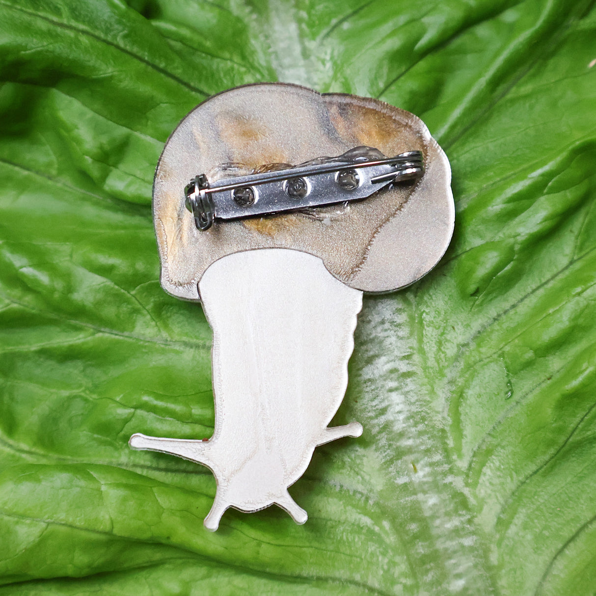 A closeup image of the back side of an acrylic snail brooch, showing the stainless steel brooch pin with rolling clasp. 