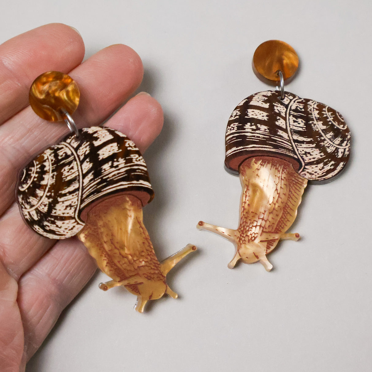 Acrylic snail-shaped earrings held in a hand with round earring toppers. 