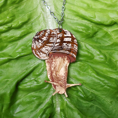 A closeup of an acrylic snail pendant necklace resting on a leaf. 