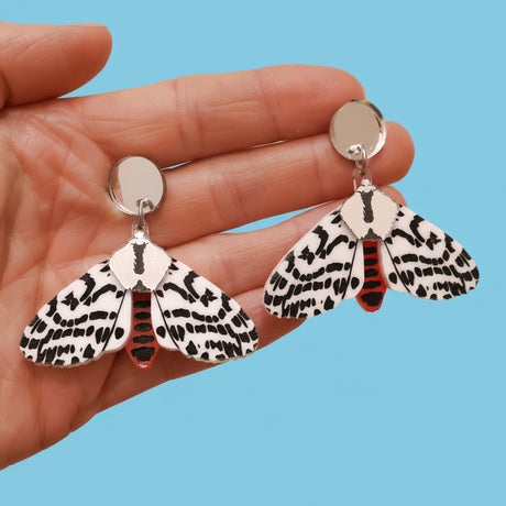 A pair of acrylic tiger moth earrings being held up by a hand. Earrings are black white and red, with silver mirror components.