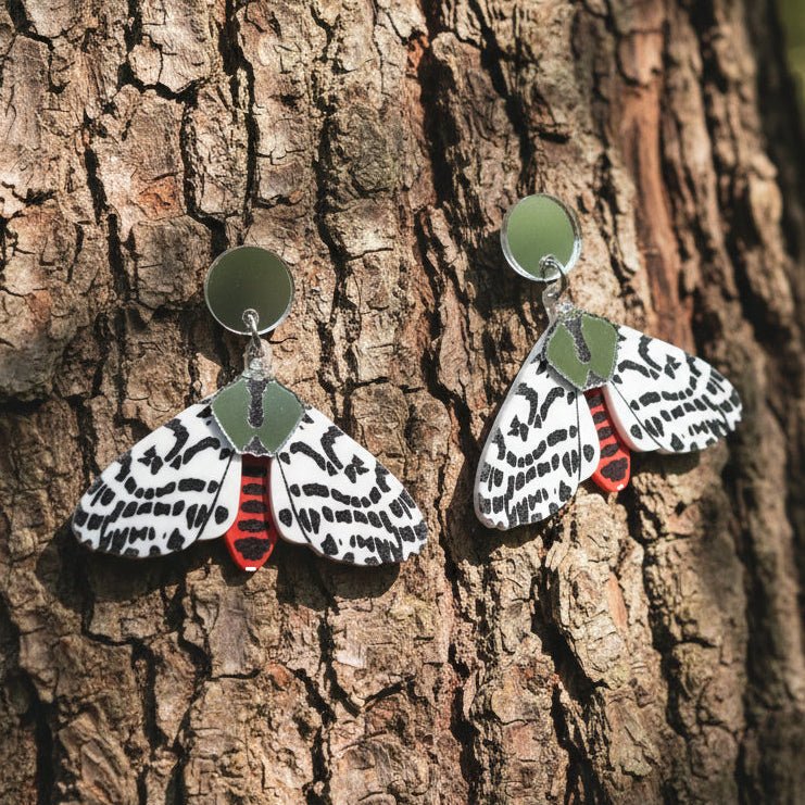 A pair of acrylic tiger moth earrings hanging against the trunk of a tree. Earrings are black white and red, with silver mirror components.