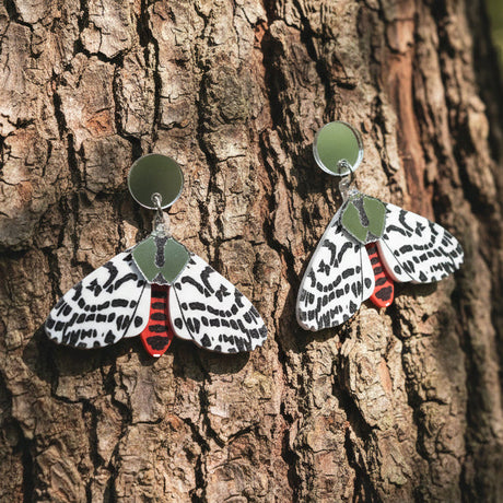 A pair of acrylic tiger moth earrings hanging against the trunk of a tree. Earrings are black white and red, with silver mirror components.
