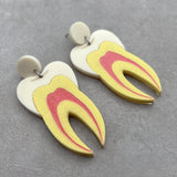 A pair of tooth earrings, laser cut from acrylic and showing a cross-sectional view. They hang from ivory toned acrylic toppers.