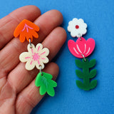 Pair of colourful asymmetrical acrylic floral earrings being held up in a hand against a blue background