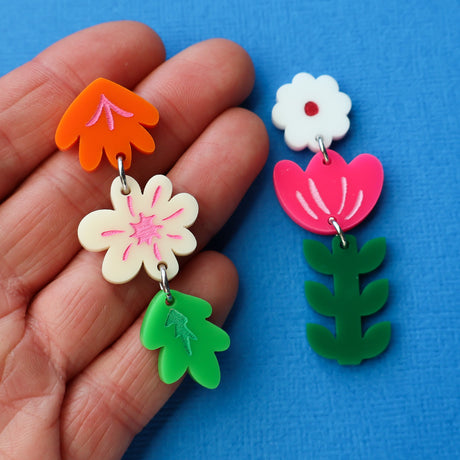 Pair of colourful asymmetrical acrylic floral earrings being held up in a hand against a blue background