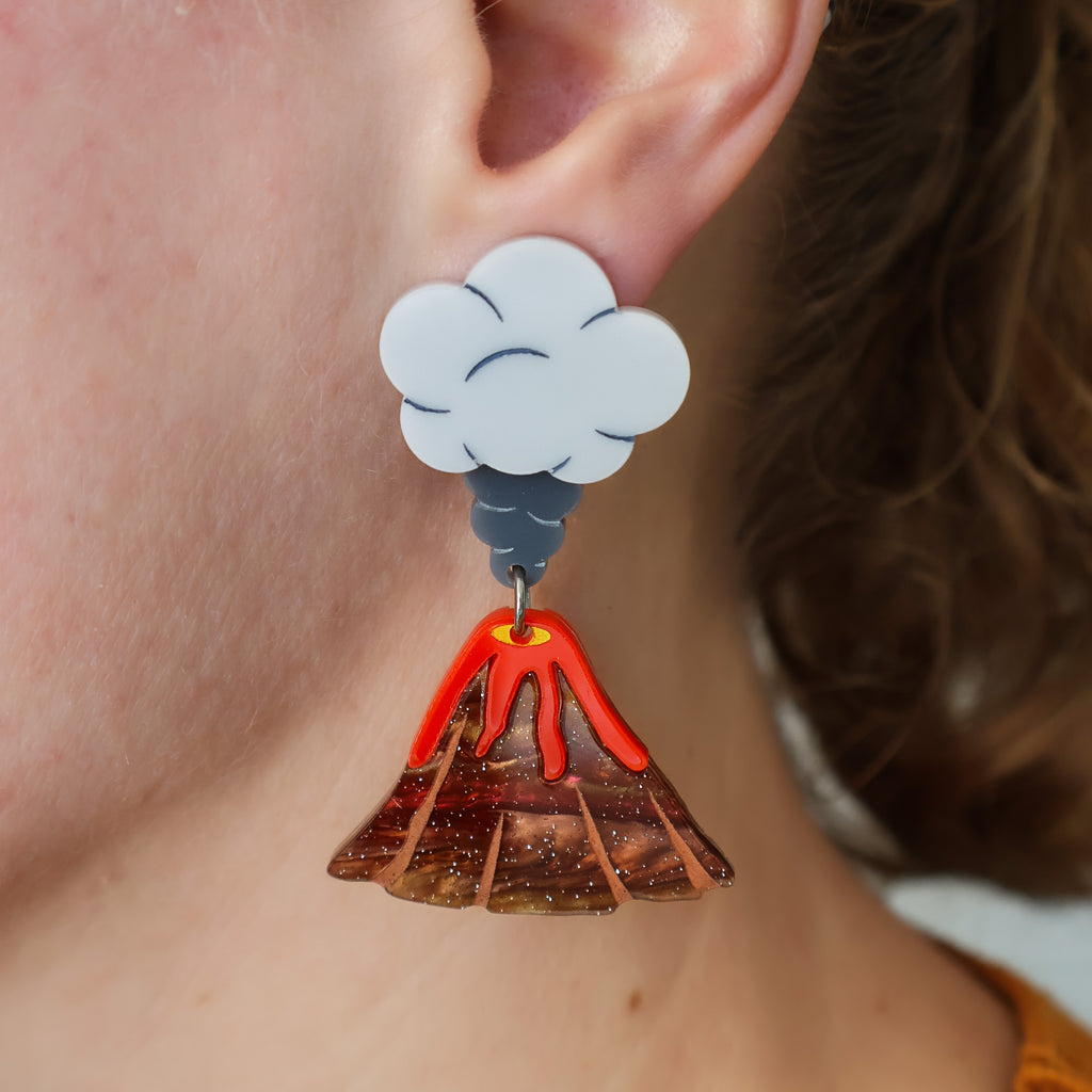 A closeup image of acrylic volcano earrings being modelled. The tops of the earrings are grey toned ash clouds, and volcanoes with lava flowing out dangle below.