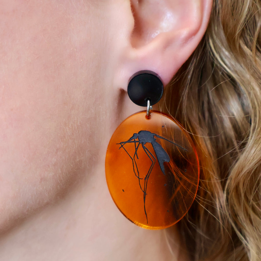 Laser cut acrylic earrings appearing like mosquitoes trapped in amber, being modelled.
