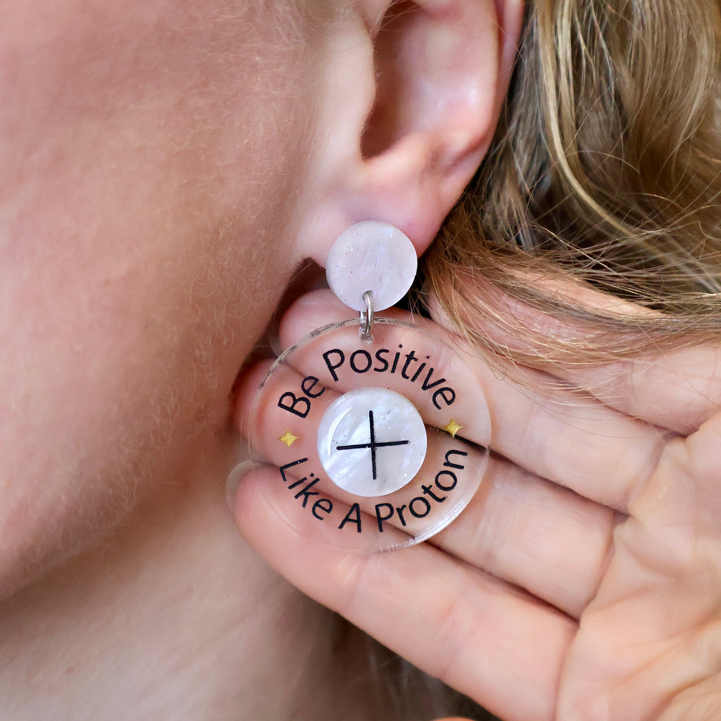 Laser cut earrings with the text 'Be Positive Like a Proton' being modelled.