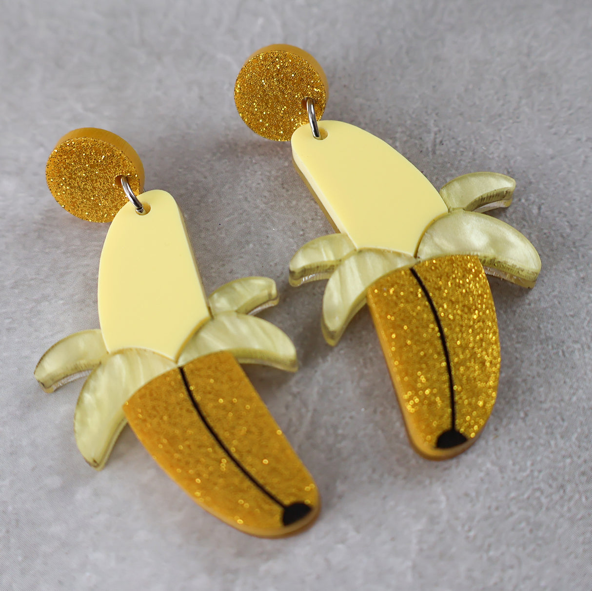 Banana-shaped earrings half peeled with yellow and gold glittery finishes on a grey background