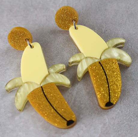 Banana-shaped earrings half peeled with yellow and gold glittery finishes on a grey background