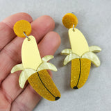 Banana-shaped earrings half peeled with yellow and gold glittery finishes held by a hand on a light grey background.