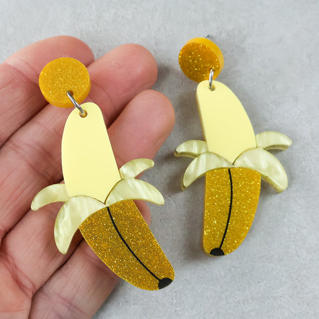 Banana-shaped earrings half peeled with yellow and gold glittery finishes held by a hand on a light grey background.