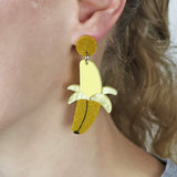 Acrylic earrings shaped like bananas being peeled worn on an ear of a woman with blonde hair. 