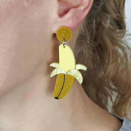 Acrylic earrings shaped like bananas being peeled worn on an ear of a woman with blonde hair. 