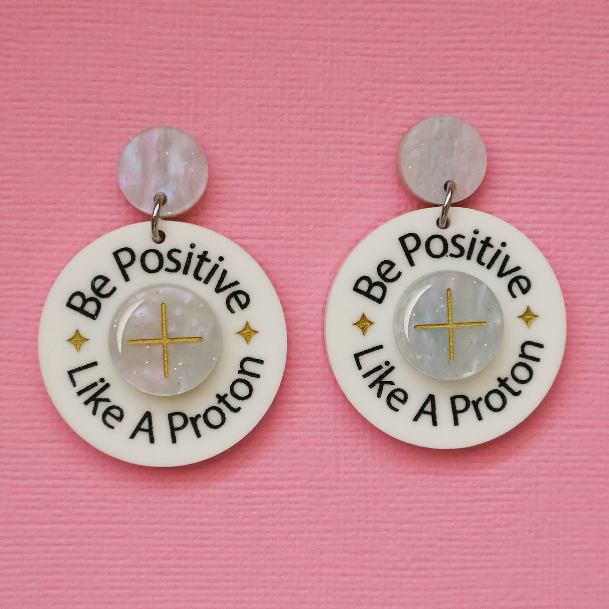 A pair of acrylic earrings with 'Be Positive Like A Proton' text on a pink background