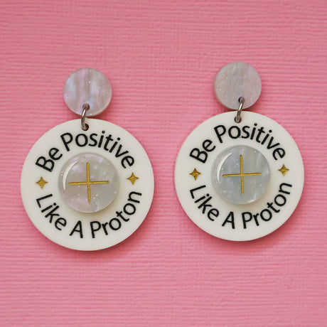 A pair of acrylic earrings with 'Be Positive Like A Proton' text on a pink background