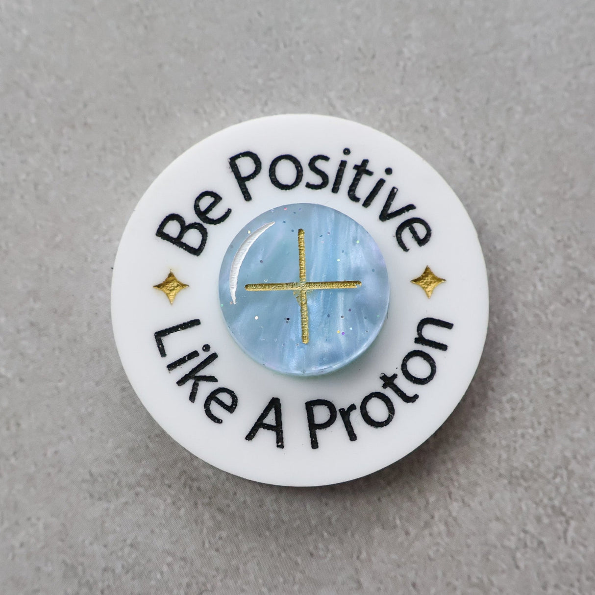 Circular brooch with 'Be Positive Like A Proton' text and a blue 'proton' in the centre
