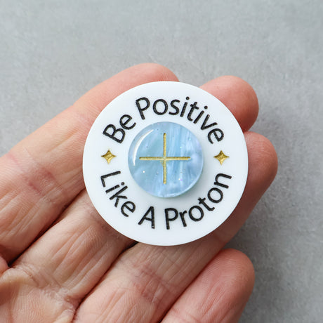 Circular brooch with 'Be Positive Like A Proton' text held in a hand