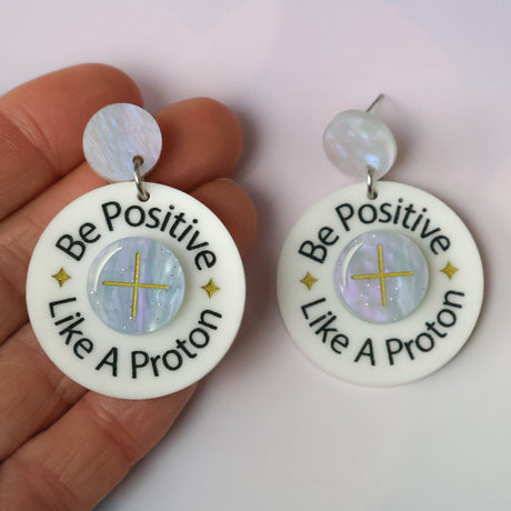 Earrings with 'Be Positive Like A Proton' text held by a hand on a light background