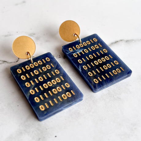 A pair of blue rectangular acrylic earrings with gold binary text engraved within, hanging from gold toned acrylic toppers. 