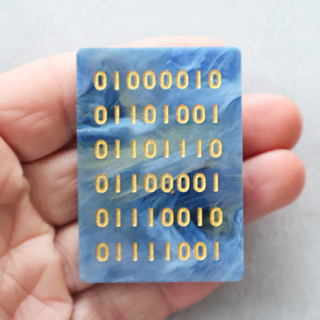 Blue rectangular acrylic brooch with binary code pattern held in a hand against