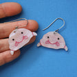 A pair of pink acrylic blobfish earrings being held in a hand.