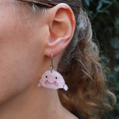 Closeup of a person wearing acrylic blobfish earrings, hanging from stainless steel french hooks. 