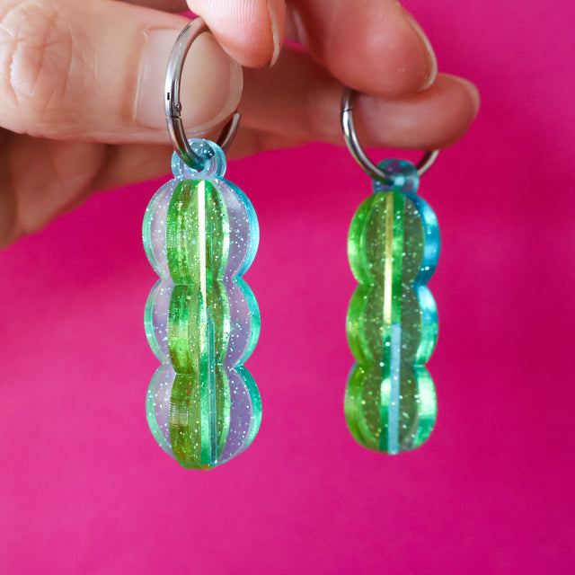 A pair of blue and green transparent acrylic earrings with a 3D construction and huggie hoops, being held up against a pink background. 