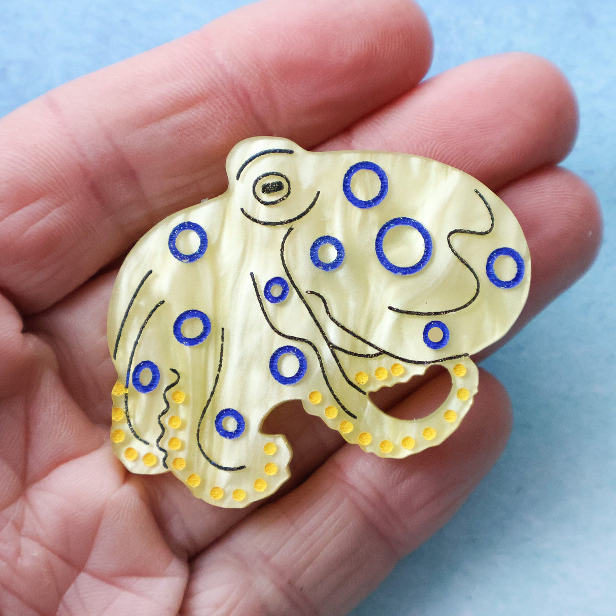 Decorative brooch shaped like an octopus with blue and yellow accents held in a hand against a light blue background.