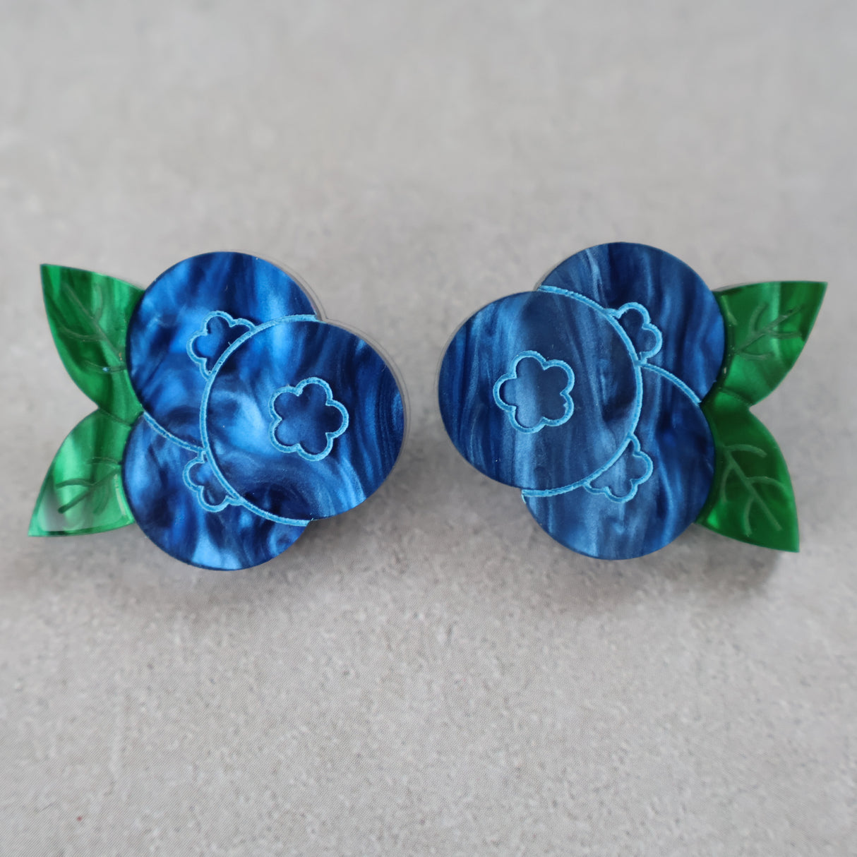 A pair of marbled acrylic blueberry studs with green leaves on a grey background