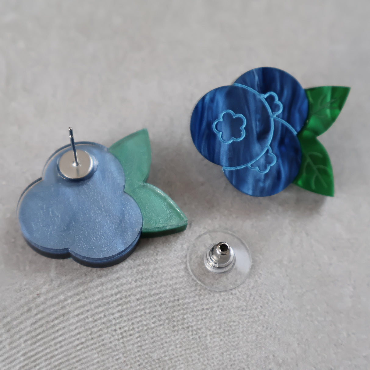 Blue and green blueberry acrylic earrings on a gray surface. One earring is turned to show the stainless steel earring post. 