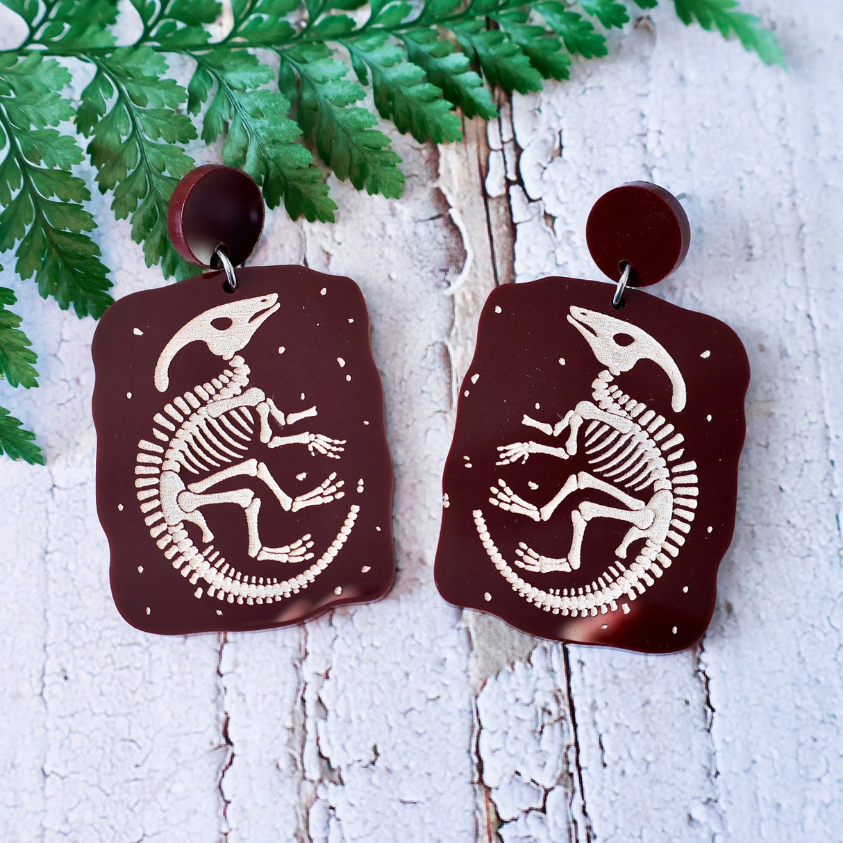 Pair of brown earrings with dinosaur skeleton design on a wooden surface with a fern nearby