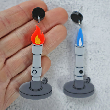 Asymmetrical Bunsen burner earrings with red and blue flame design held by a hand against a textured background.