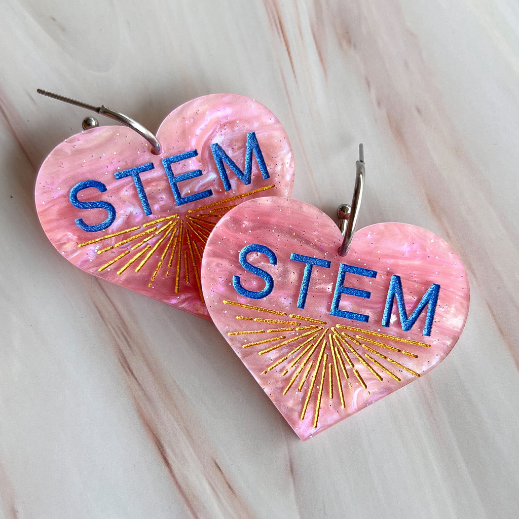 Swirly pink acrylic heart earrings with blue STEM text and a gold starburst pattern, hanging from stainless steel toppers. Closeup view. 