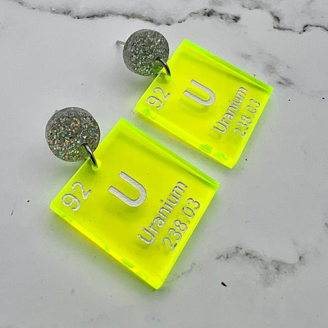 Uranium periodic table earrings, with white text on bright yellow transparent acrylic squares, hanging from silver glitter toppers.