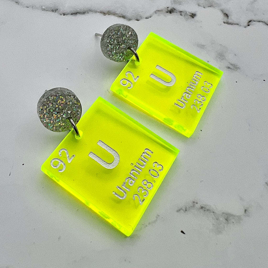 Uranium periodic table earrings, with white text on bright yellow transparent acrylic squares, hanging from silver glitter toppers.