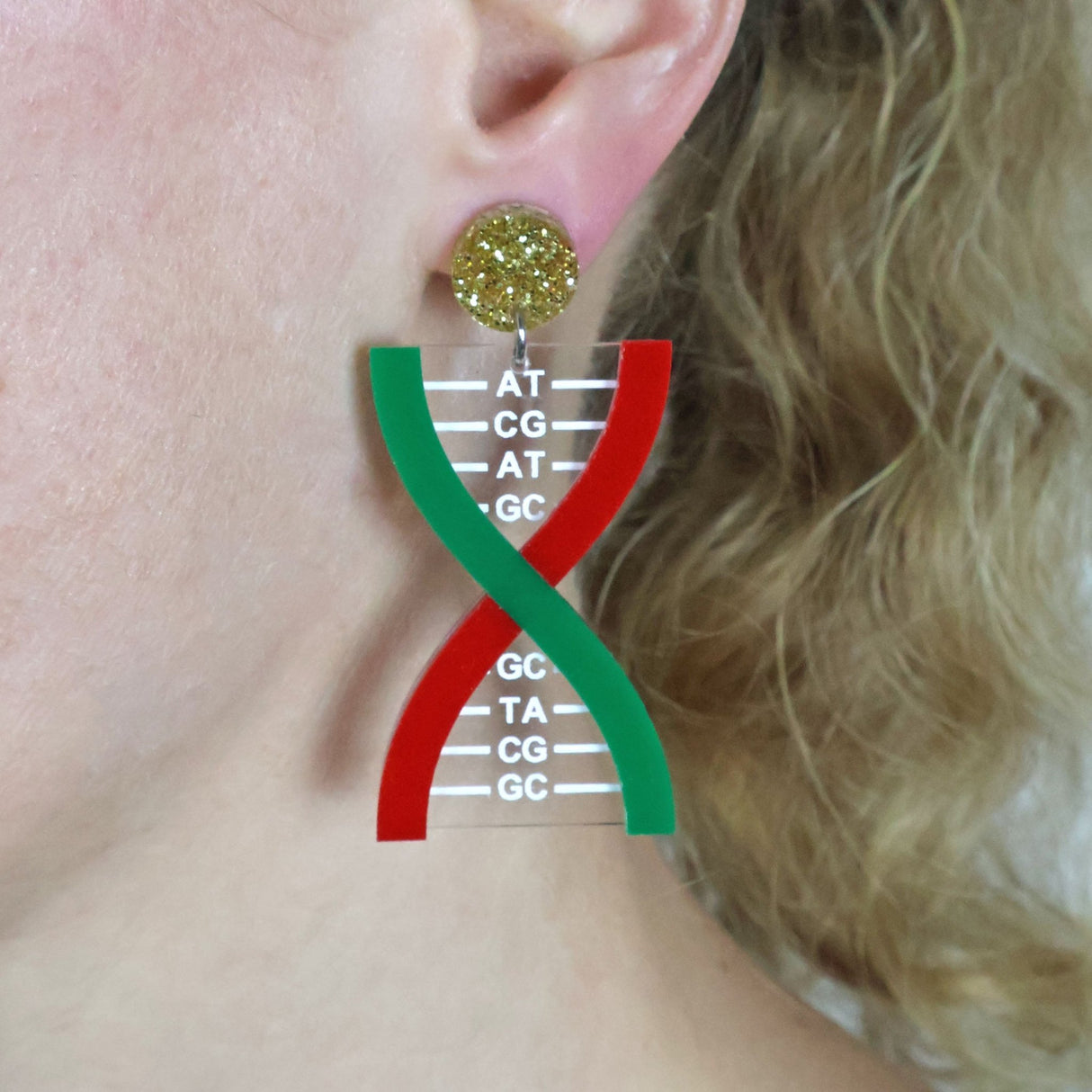 Acrylic Earrings shaped like DNA helixes with red and green design and visible base pairs, and gold glittery acrylic toppers, worn by a person.