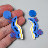 A pair of blue and white chromodoris sea slug earrings being held in a hand